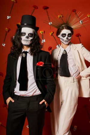 Foto de Pareja en día de los muertos maquillaje y traje festivo posando sobre fondo rojo con claveles - Imagen libre de derechos