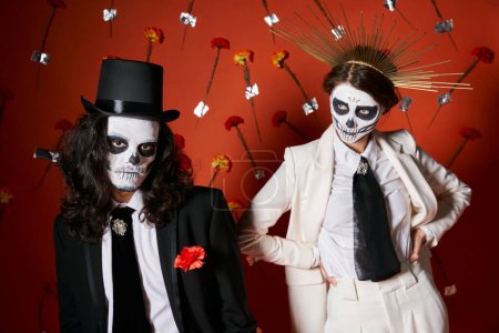 Foto de Pareja en día de los muertos cráneo maquillaje y elegante atuendo posando sobre fondo rojo con claveles - Imagen libre de derechos