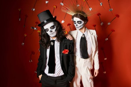 elegant couple in dia de los muertos skull makeup looking at camera on red backdrop with flowers