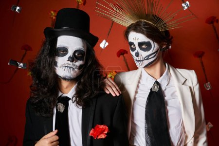 Photo for Trendy couple in dia de los muertos skull makeup looking at camera on red backdrop with carnations - Royalty Free Image