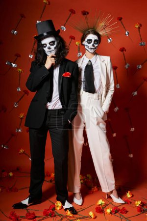 full length of festive elegant couple in dia de los muertos makeup on red backdrop with floral decor