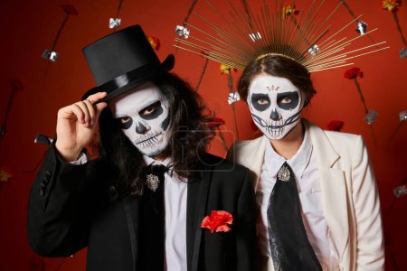 couple in dia de los muertos makeup,  man touching top hat near woman in crown, red floral backdrop