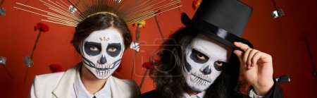 Photo for Man in skull makeup touching top hat near woman in crown on red background with flowers, banner - Royalty Free Image