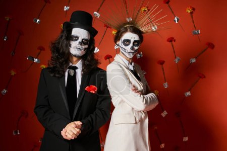 woman in crown and skull makeup with folded arms near spooky man in top hat on red floral backdrop