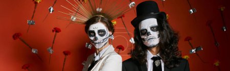 couple in dia de los muertos makeup and festive attire posing on red backdrop with flowers, banner