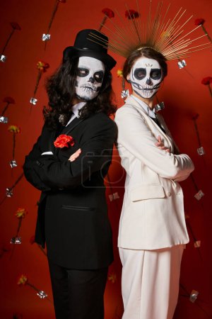 Elegantes Paar in Zuckerschädel-Make-up, Rücken an Rücken stehend mit verschränkten Armen vor rotem Blumenhintergrund