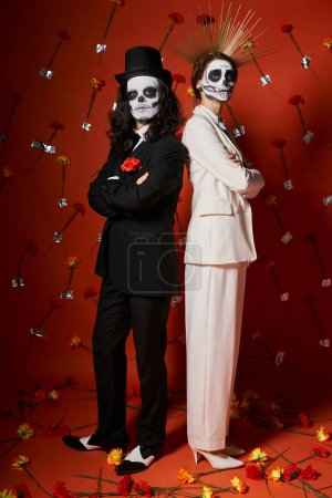 Téléchargez les photos : Couple branché dans le maquillage catrina debout dos à dos avec les bras croisés sur fond rouge avec des fleurs - en image libre de droit
