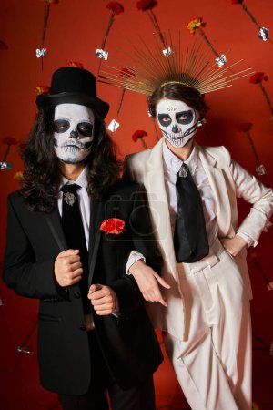 woman in skeleton makeup and crown looking at camera near man in top hat on red floral backdrop