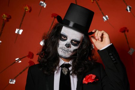 extravaganter Mann in Totenkopf-Make-up, der Zylinder auf rotem Hintergrund mit Nelken berührt, Tag des Todes