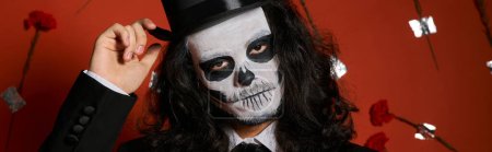 extravagant man in Day of Dead makeup touching top hat on red backdrop with carnations, banner magic mug #676495928