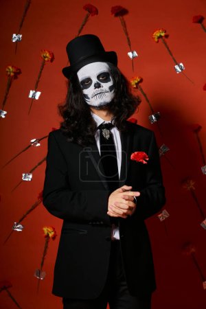 man in skull makeup and festive attire looking at camera on red floral backdrop, dia de los muertos