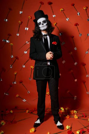 full length of man in skull makeup and festive attire on red floral backdrop, dia de los muertos