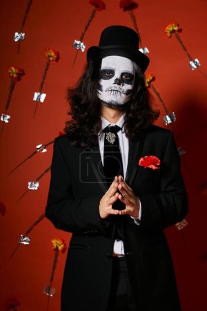 elegant man in dia de los muertos skull makeup looking at camera in red studio with carnations
