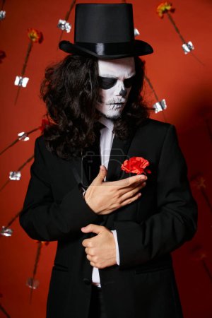elegant man in dia de los muertos skull makeup touching carnation on blazer on red floral backdrop