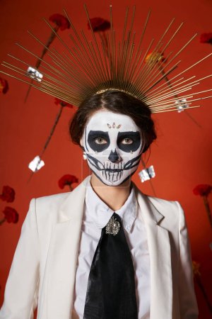 extravagant woman in skull makeup and white suit with crown looking at camera on red floral backdrop