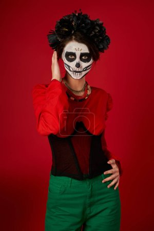woman in traditional dia de los muertos skull makeup and festive attire looking at camera on red