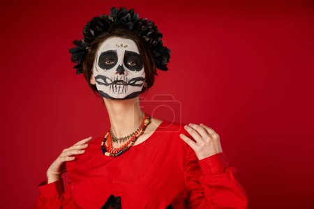 Photo for Woman in traditional dia de los muertos skull makeup and colorful beads looking at camera on red - Royalty Free Image