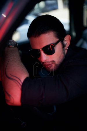 sexy joven conductor con tatuaje y gafas de sol posando en el volante del coche mirando a la cámara
