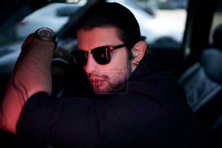 gut aussehender Mann mit Tätowierung und schwarzem Hemd posiert hinter dem Lenkrad seines Autos