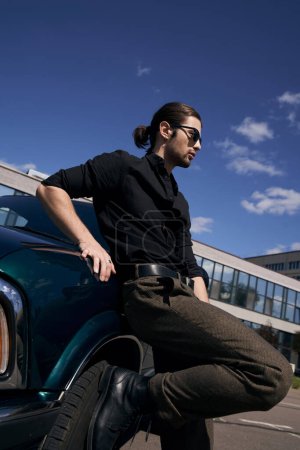 sexy hombre elegante con barba y cola de caballo posando cerca de su coche verde y mirando hacia otro lado, concepto de moda