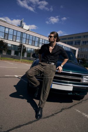 joven hombre guapo con barba y cola de caballo en traje negro posando cerca de coche, concepto de moda