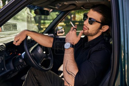 Photo for Good looking bearded man with ponytail and sunglasses smoking cigarette while posing in his car - Royalty Free Image