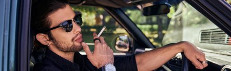 young bearded man with wristwatch and sunglasses smoking cigarette while posing in car, banner