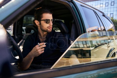 atractivo hombre elegante con cola de caballo y gafas de sol en traje casual negro fumar cigarrillo en el coche