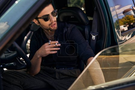 sexy modelo masculino en camisa negra con gafas de sol detrás del volante de conducción y fumar cigarrillo