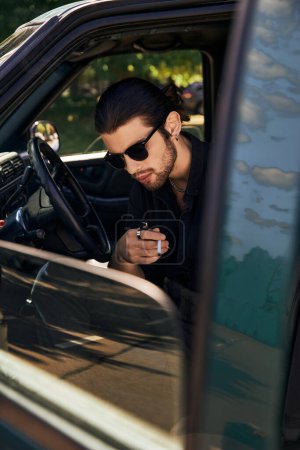schöner sexy Mann mit Sonnenbrille und Pferdeschwanz entspannt hinter dem Lenkrad mit Zigarette in der Hand