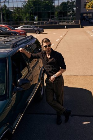 tiro vertical de hombre atractivo en traje negro con accesorios posando cerca de su coche, concepto de moda