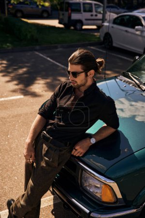stylischer, attraktiver Mann mit Sonnenbrille im schwarzen Outfit, an Auto gelehnt und wegschauend, sexy Fahrer