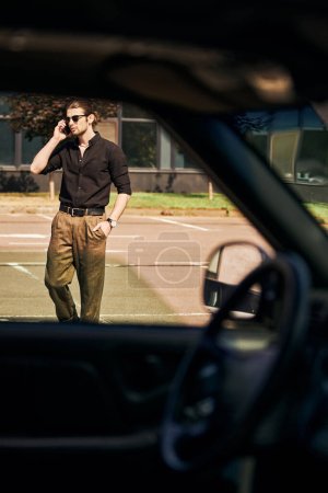 good looking man with dapper look with hand in pocket talking by phone with car insurer, sexy driver