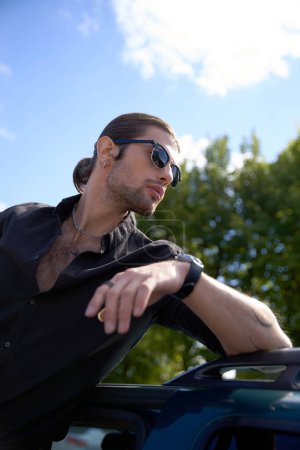 vertikale Aufnahme eines sexy jungen Fahrers mit Accessoires im schwarzen Outfit, der mit Ellbogen auf seinem Auto posiert