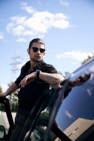 beau jeune modèle masculin avec des lunettes de soleil et tatouage posant en plein air avec le bras sur sa voiture, chauffeur sexy