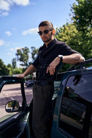 auffälliger junger Mann in stylischem schwarzen Outfit, der neben seinem Auto steht und in die Kamera schaut, Mode