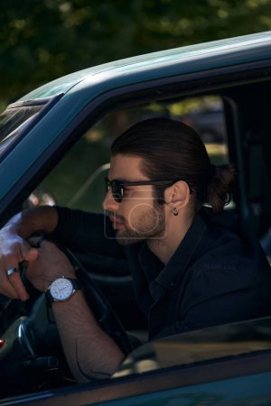 Foto de Hombre elegante de buen aspecto con pendiente y barba en traje negro detrás del volante, conductor sexy - Imagen libre de derechos
