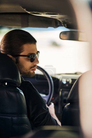 vertical shot of good looking male model in black elegant attire at steering wheel, turning his head