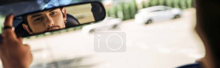 Foto de Encantador joven sentado detrás del volante y mirando en el espejo retrovisor, conductor sexy, pancarta - Imagen libre de derechos