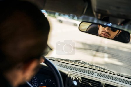 guapo sexy modelo masculino con gafas de sol detrás del volante y mirando en espejo retrovisor