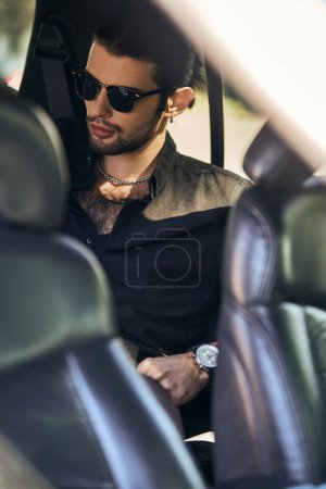 Foto de Guapo modelo masculino con mirada elegante en gafas de sol sentado en el asiento trasero de su coche, la moda y el estilo - Imagen libre de derechos
