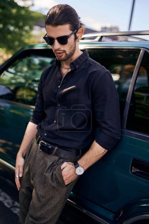 hombre sexy con estilo en camisa negra con gafas de sol posando junto a su coche al aire libre, la moda y el estilo