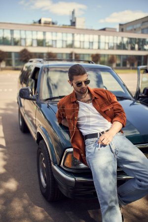 hombre sexy de buen aspecto en traje brillante de moda posando junto a su coche con el cigarrillo en la mano