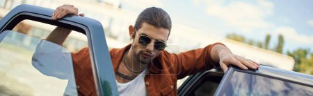 Photo for Good looking man with sunglasses and ponytail in vibrant outfit posing next to his car, banner - Royalty Free Image