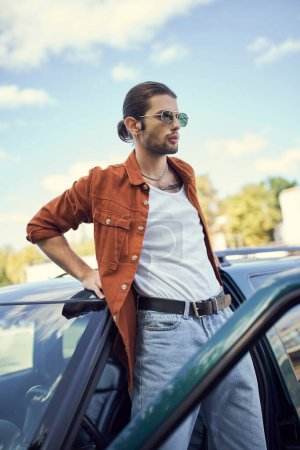 encantador modelo masculino joven en camisa marrón y vaqueros azules de pie junto a su coche y mirando hacia otro lado