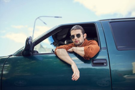 guapo joven modelo masculino con gafas de sol mirando a través de la ventana del coche en la cámara, moda y estilo