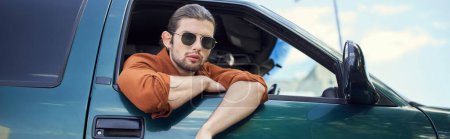 atractivo hombre elegante con gafas de sol mirando a través de la ventana del coche en la cámara, concepto de moda, pancarta