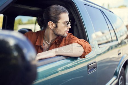 schöner Mann in stilvollem, lebendigem Outfit mit Sonnenbrille und Pferdeschwanz, der aus dem Autofenster schaut, Stil