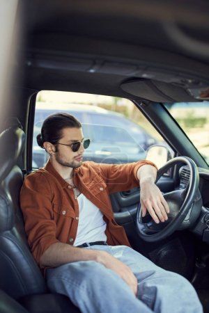 charming man with ponytail and sunglasses relaxing behind steering wheel and looking away, fashion
