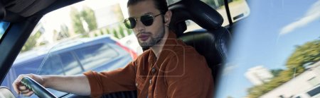 young handsome man with stylish in vivid attire posing in car and looking at camera, banner